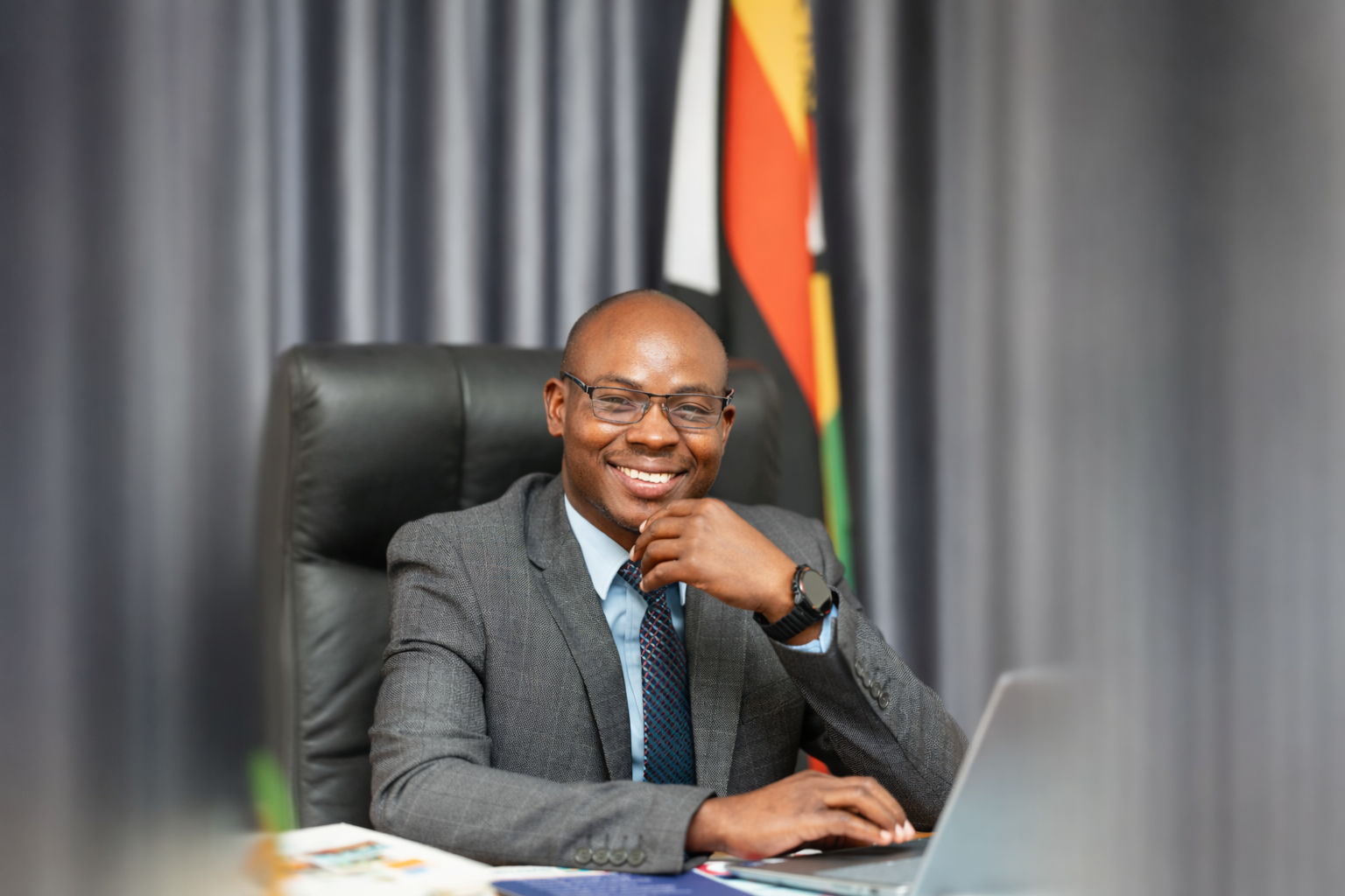A smiling man in a grey suit and glasses sits at a desk using a laptop, with a Zimbabwean flag and grey curtains in the background.