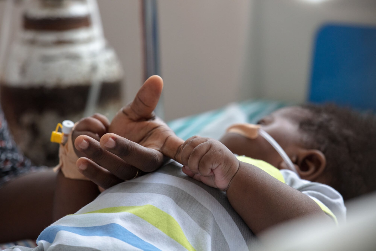 A newborn baby lying in a hospital bed gently holding their mother’s finger, symbolising vulnerability and early childhood care