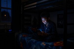 A young Black boy in blue pyjamas sits on a Superman-themed bunk bed at night while texting on his phone, illuminated by the phone screen glow.
